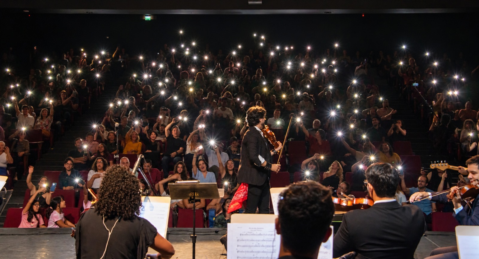 Violinista e guitarrista em palco com luz quente e demais músicos ao fundo