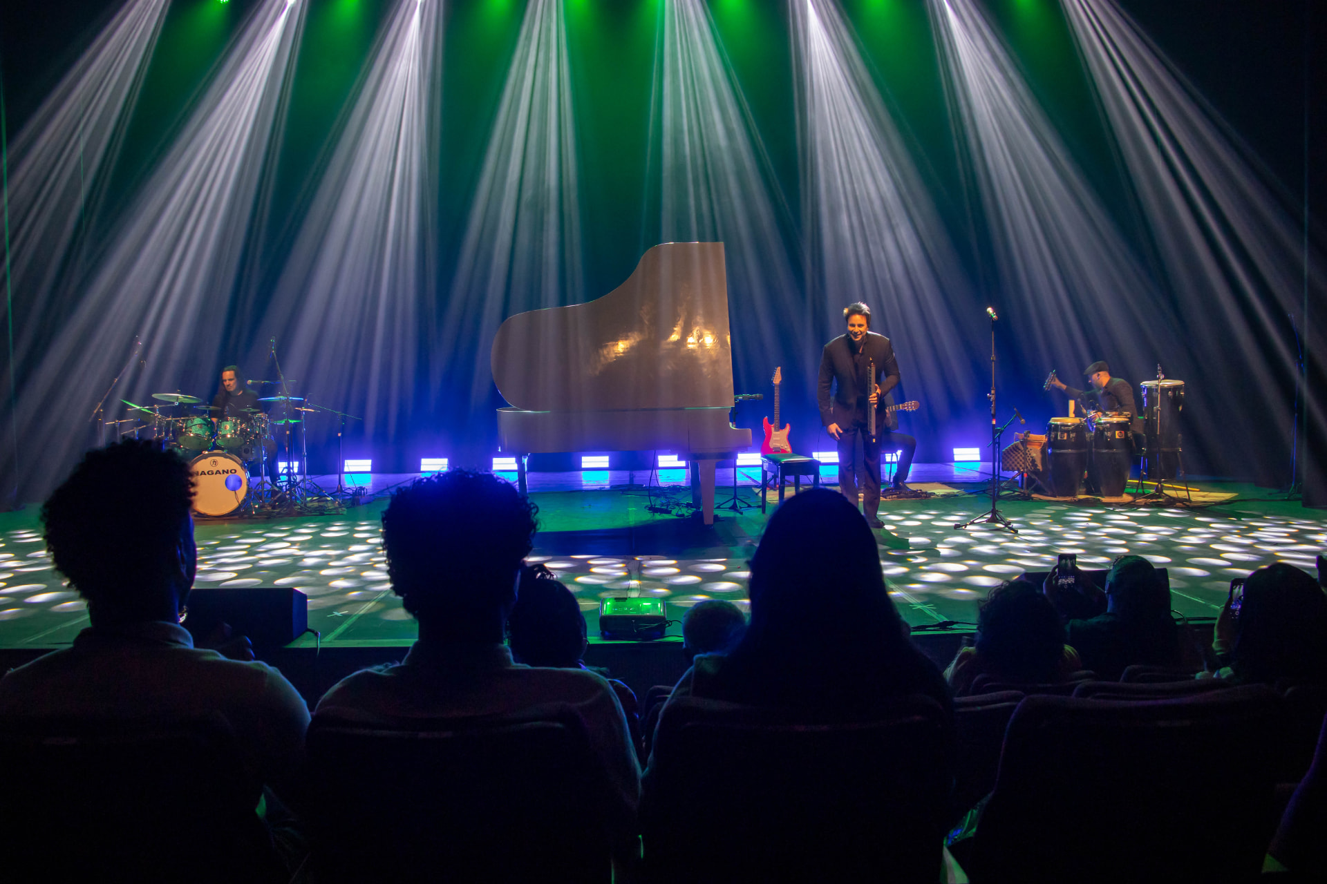 Palco com piano branco, baterista e percussionista, vistos da plateia