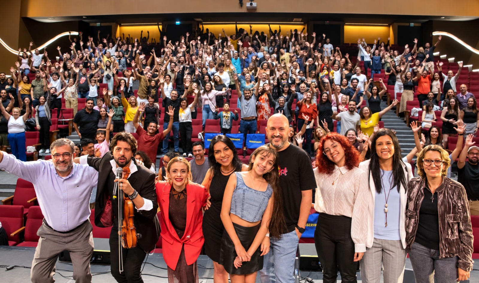 Grupo de artistas e organizadores no palco com a plateia de mãos levantadas atrás