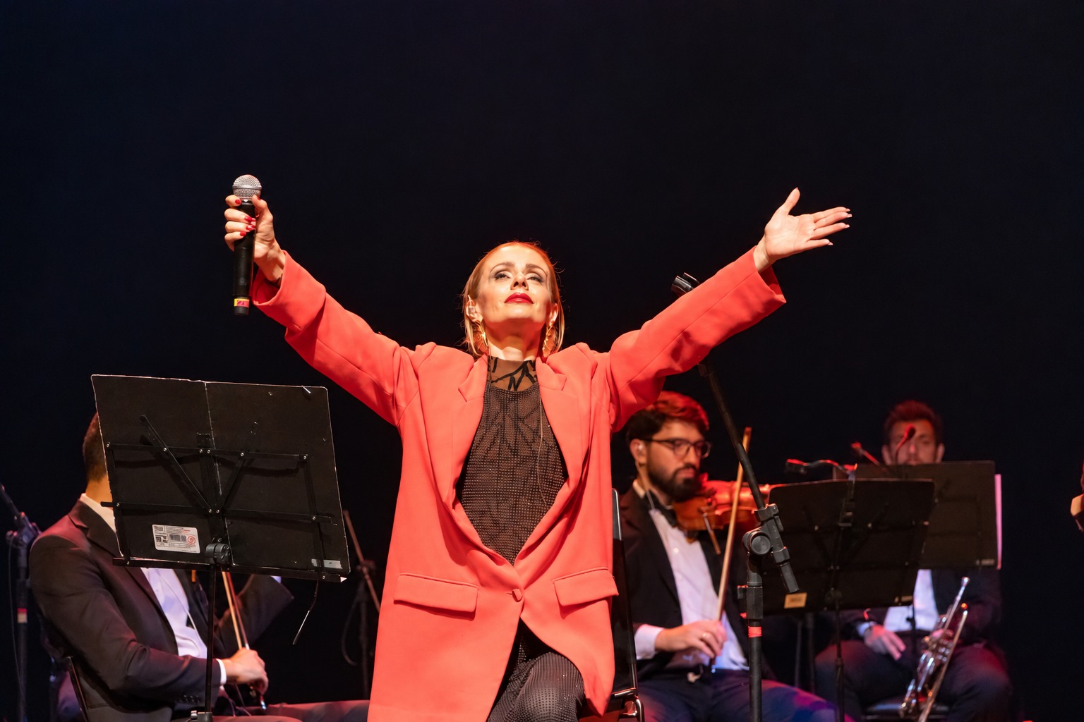 Cantora de blazer vermelho com os braços abertos e músicos de orquestra sentados ao fundo