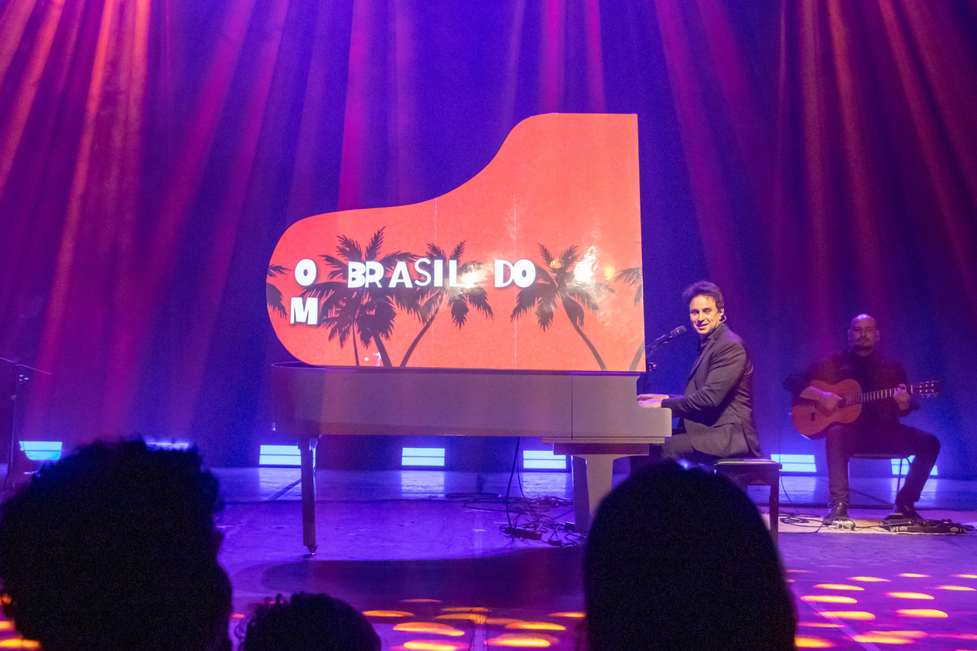 Pianista ao piano de cauda branco em palco iluminado, com plateia em silhueta na frente