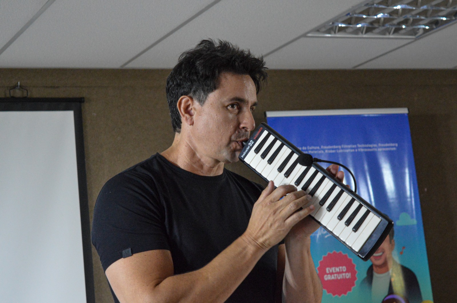 Músico tocando melódica em uma sala de aula, com banner do projeto ao fundo