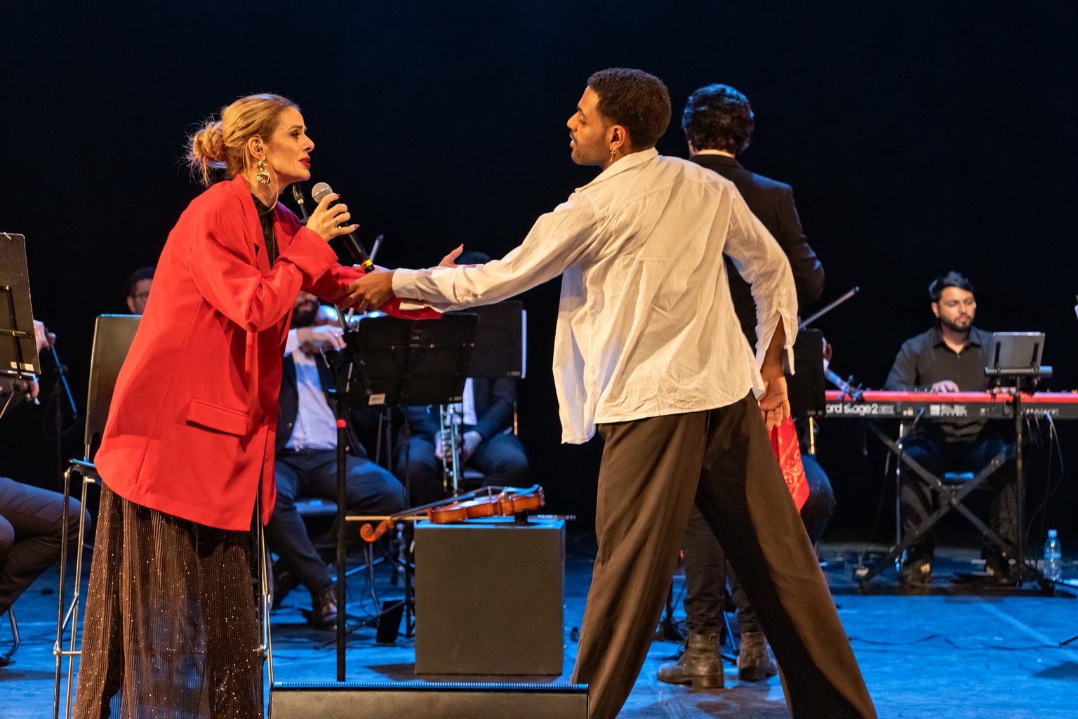 Cantora com microfone e bailarino de camisa branca em palco com luz azul